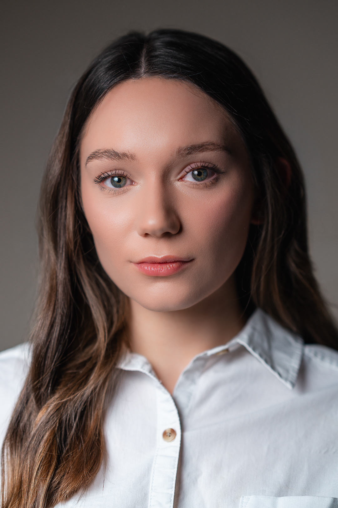 Portrait Photography | A young woman with fair skin and long brown hair looks directly at the camera. She wears a white button-up shirt and has a neutral expression. The background is plain and softly lit, drawing attention to her face. Nathan Mountain | Hampshire & London Freelance Photographer | Hampshire & London Camera Man