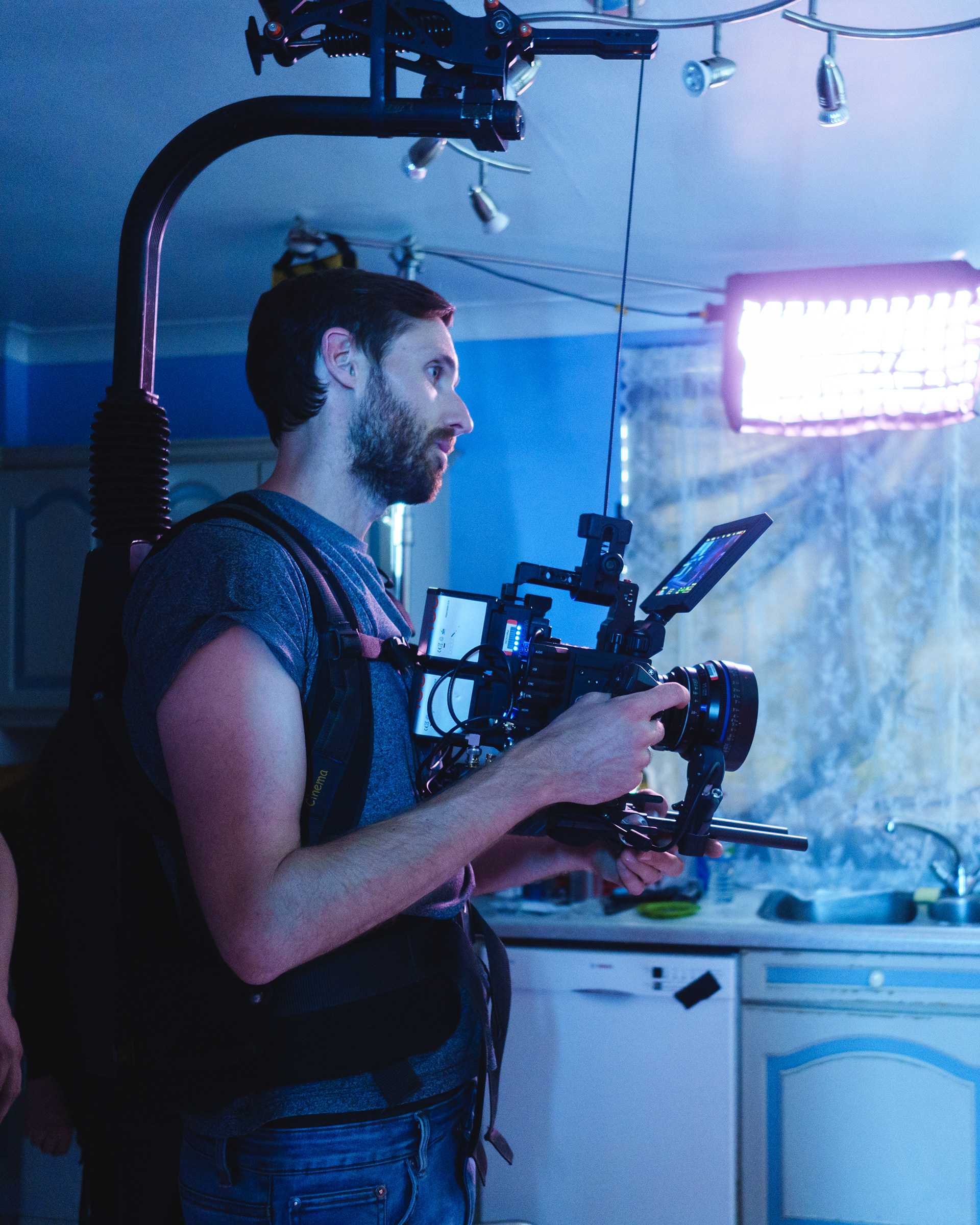 Behind The Scenes (BTS) Photography | A man operates a professional video camera with a shoulder rig and stabiliser in a kitchen. He wears a harness for support, and a bright light illuminates the scene. Behind him are kitchen cupboards, a sink, and a curtained window. Nathan Mountain | Hampshire & London Freelance Photographer | Hampshire & London Camera Man