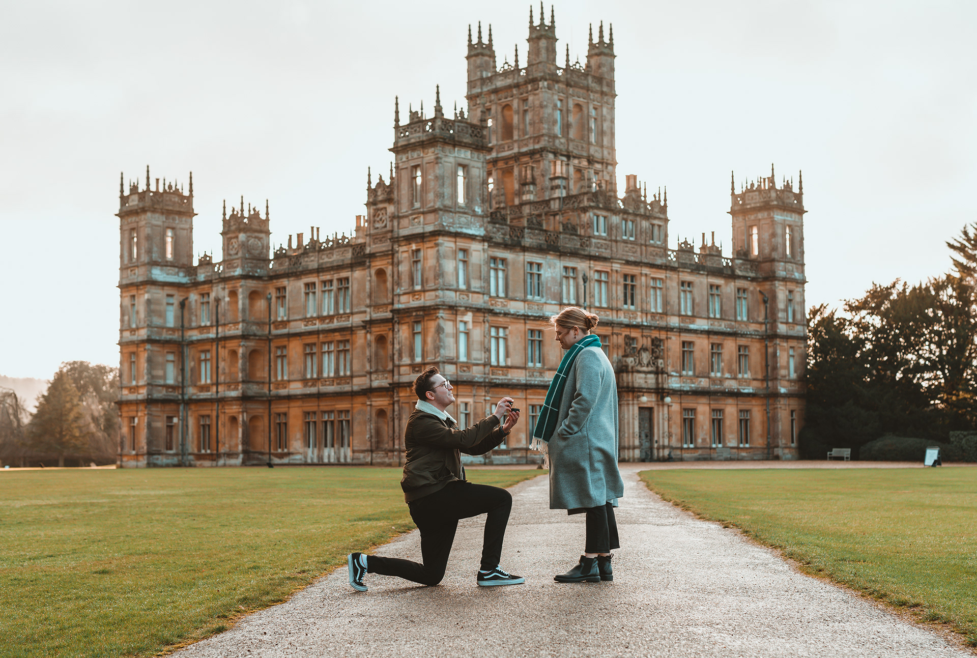 Engagement Photography | A man kneels, proposing to a woman on a wide path in front of a grand, historic building with multiple towers and ornate architecture. The woman, wearing a teal coat, looks down at him, surprised and smiling. The lawn and building suggest a formal, elegant setting. Nathan Mountain | Hampshire & London Freelance Photographer | Hampshire & London Camera Man