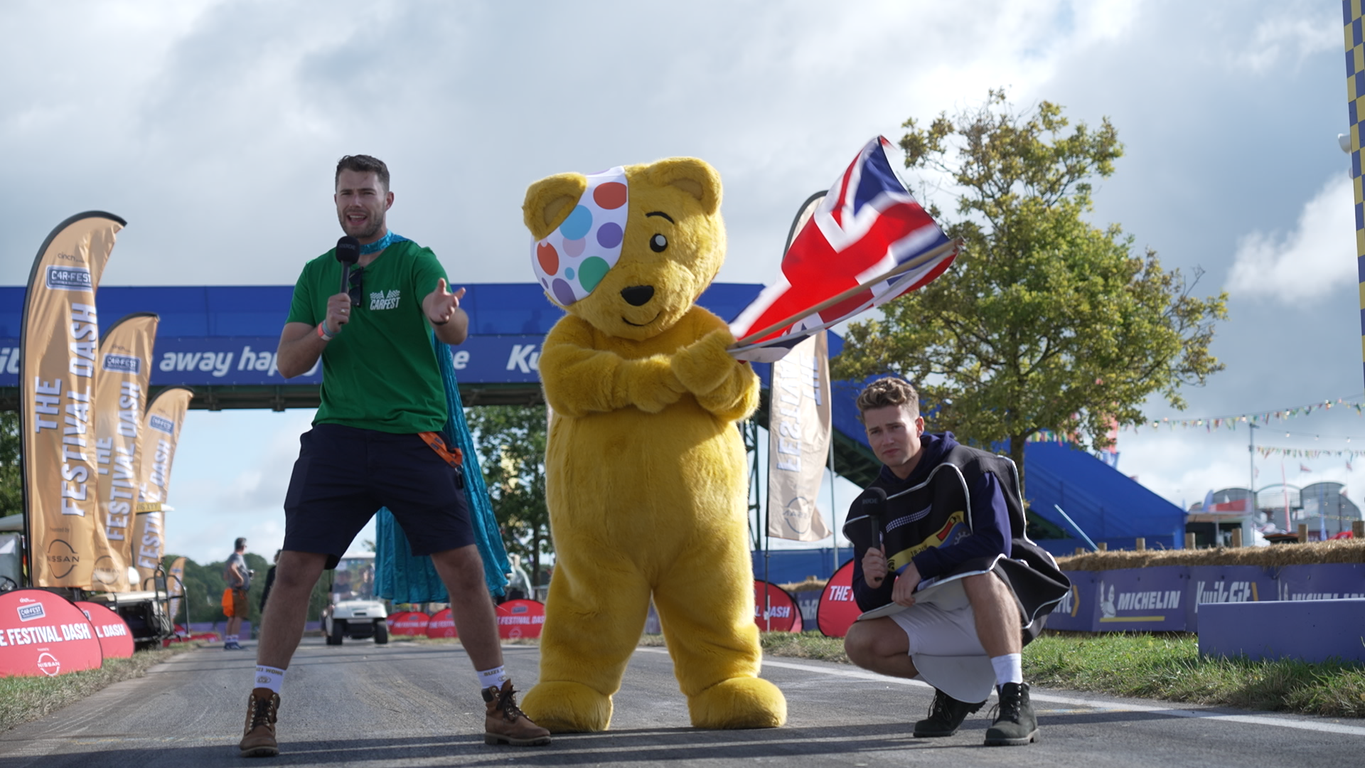 Event Photography | Three people pose at an outdoor event: one in a green shirt with a microphone, one in a yellow bear costume with a colourful spotted eye patch, and one kneeling in a black jacket holding a large British flag. Event banners and trees are visible behind them. Nathan Mountain | Hampshire & London Freelance Photographer | Hampshire & London Camera Man