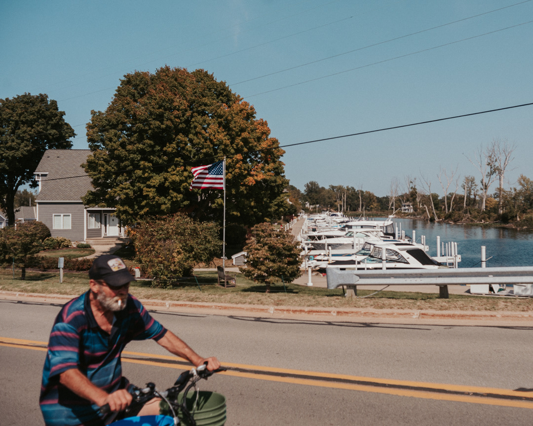 Personal Photography | An older man with a grey beard, wearing a cap and striped shirt, rides a bicycle along a road. Behind him, an American flag flies in front of a house by a marina with several docked boats on a sunny day. Trees line the waterfront. Nathan Mountain | Hampshire & London Freelance Photographer | Hampshire & London Camera Man