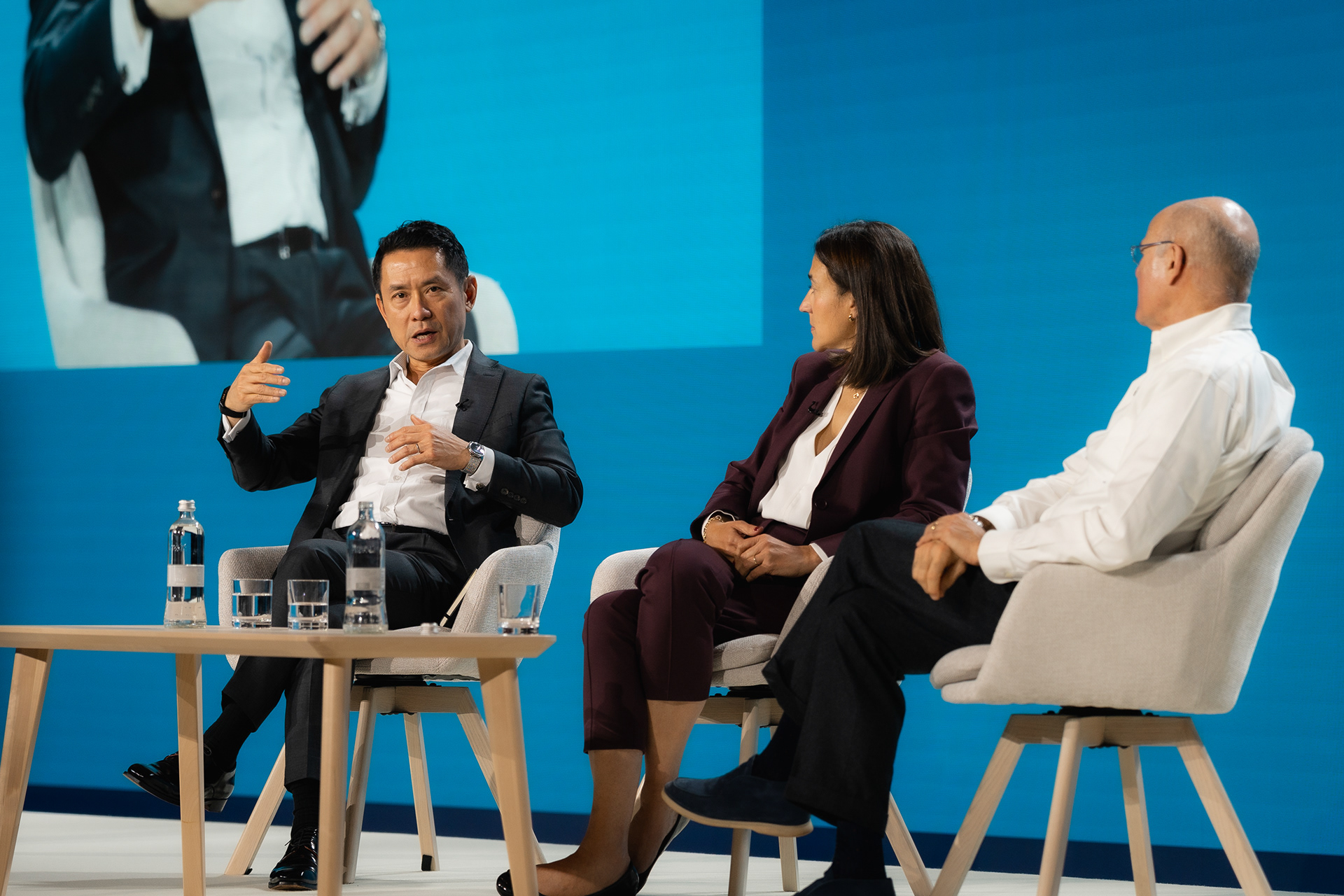Event Photography | Three people sit on stage in modern chairs, having a panel discussion. The man on the left gestures while speaking, the woman in the centre listens attentively, and the man on the right sits relaxed. A blue backdrop and water bottles are visible on the table in front of them. Nathan Mountain | Hampshire & London Freelance Photographer | Hampshire & London Camera Man