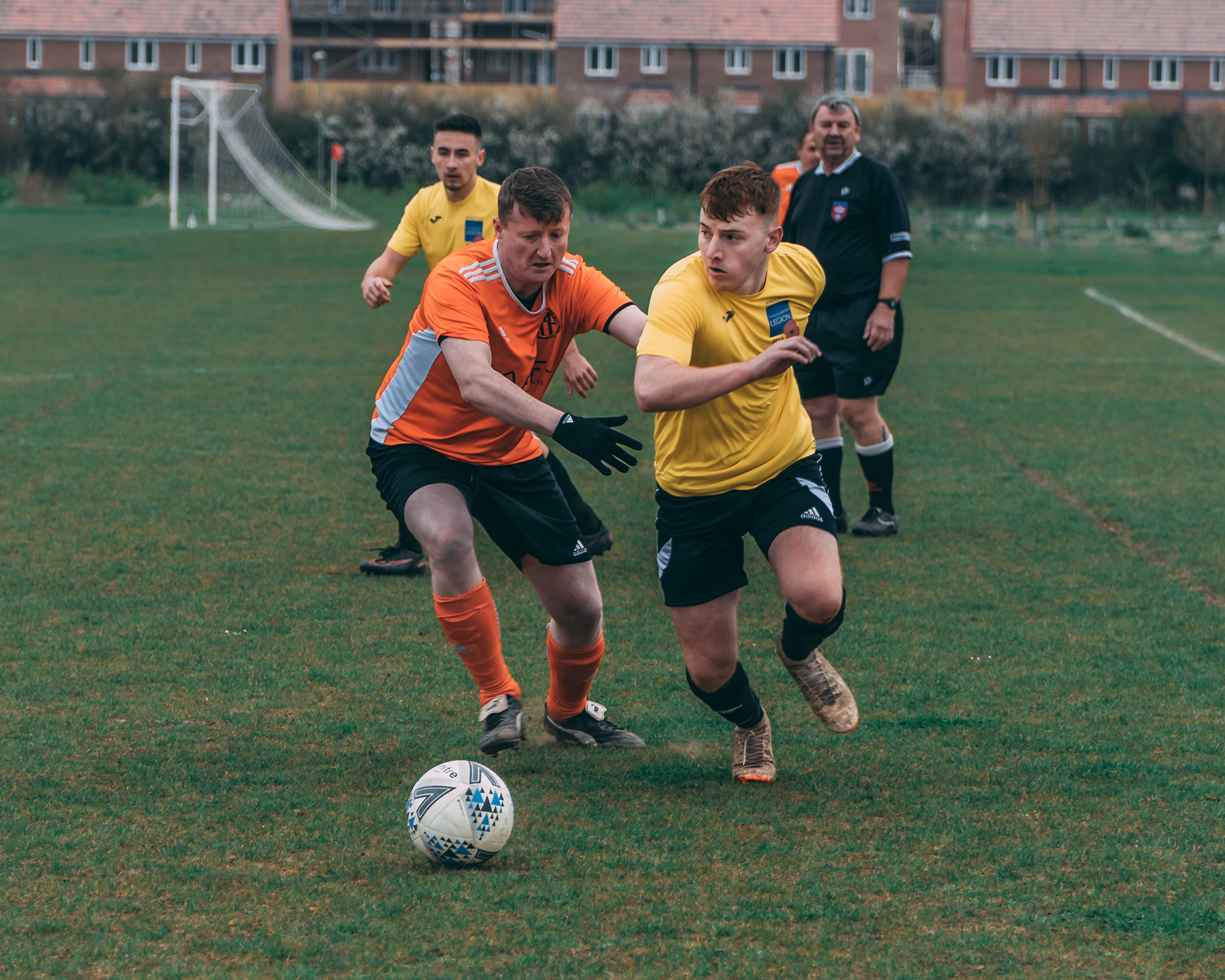 Sports & Fitness Photography | Two young men compete for the ball during a football match; one in an orange kit, the other in yellow, both focused and running. A referee and two other players are seen in the background on a grassy pitch with a goal and houses visible. Nathan Mountain | Hampshire & London Photography | Hampshire & London Camera Man