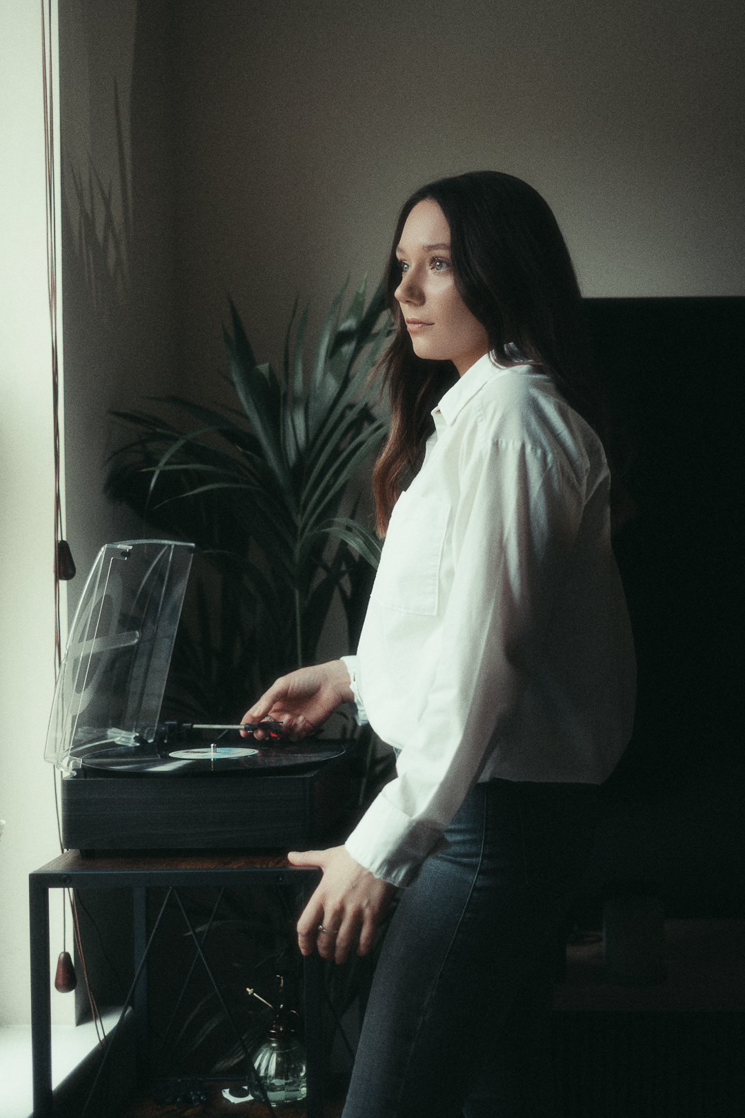 Fashion Photography | A woman with long brown hair, wearing a white shirt and jeans, stands indoors beside a record player. She looks out of a window, soft light illuminating her face. A potted plant and furniture are visible in the background, creating a cosy, relaxed atmosphere. Nathan Mountain | Hampshire & London Freelance Photographer | Hampshire & London Camera Man