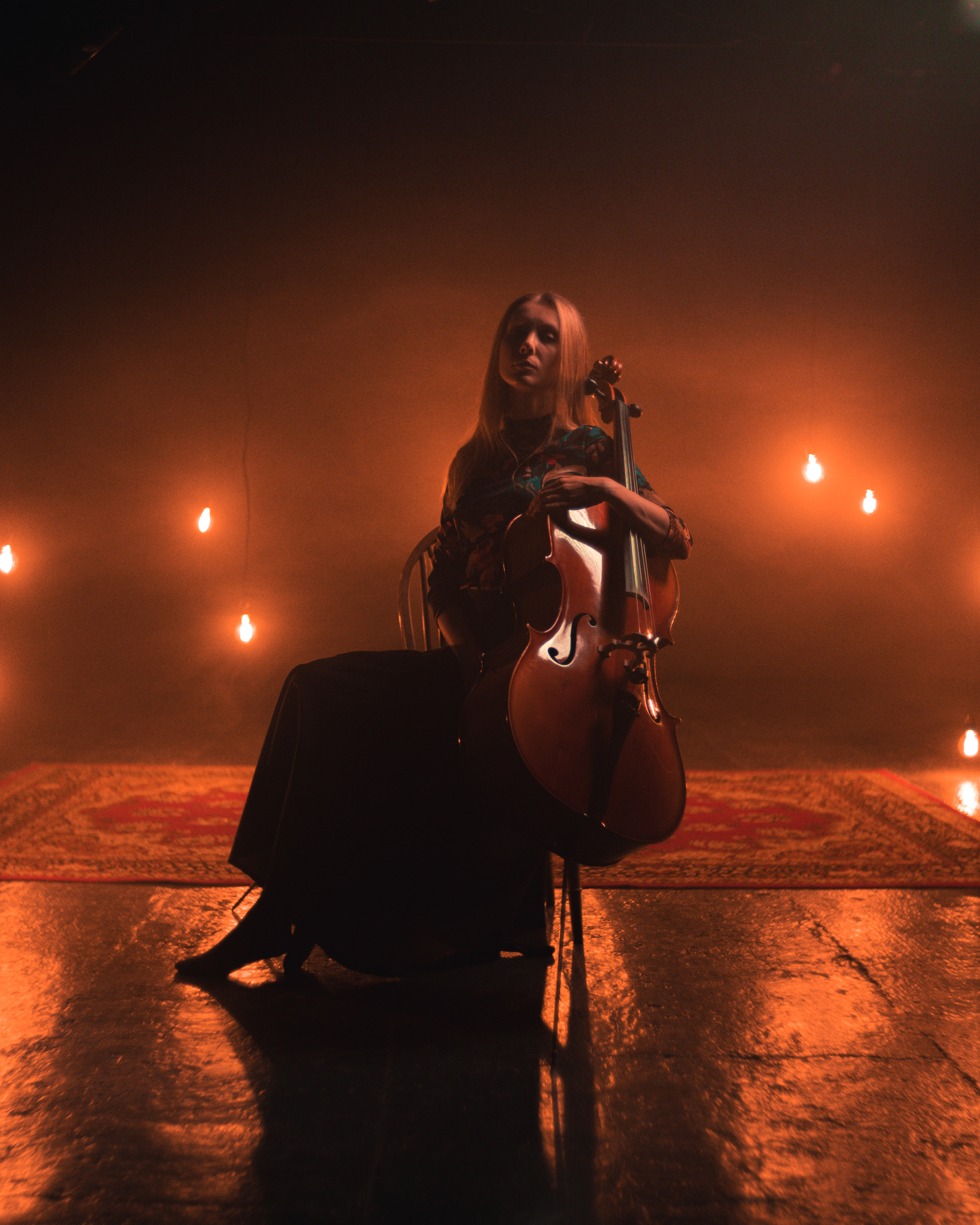 Music Photography | A woman sits on a chair holding a cello in a dimly lit room. Warm, hanging light bulbs and an ornate red carpet create a dramatic, atmospheric scene. She wears a long dark dress and is lit softly from the front, with shadows behind her. Nathan Mountain | Hampshire & London Freelance Photographer | Hampshire & London Camera Man