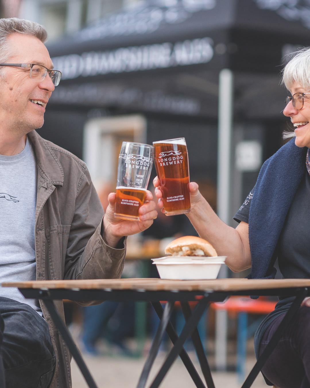 Commercial Photography | Two smiling adults sit at an outdoor table, clinking pint glasses with amber beer. A small tray with a sandwich or burger is on the table. They appear relaxed and happy, with brewery marquees and blurred background suggesting a festive outdoor event. Nathan Mountain | Hampshire & London Freelance Photographer | Hampshire & London Camera Man