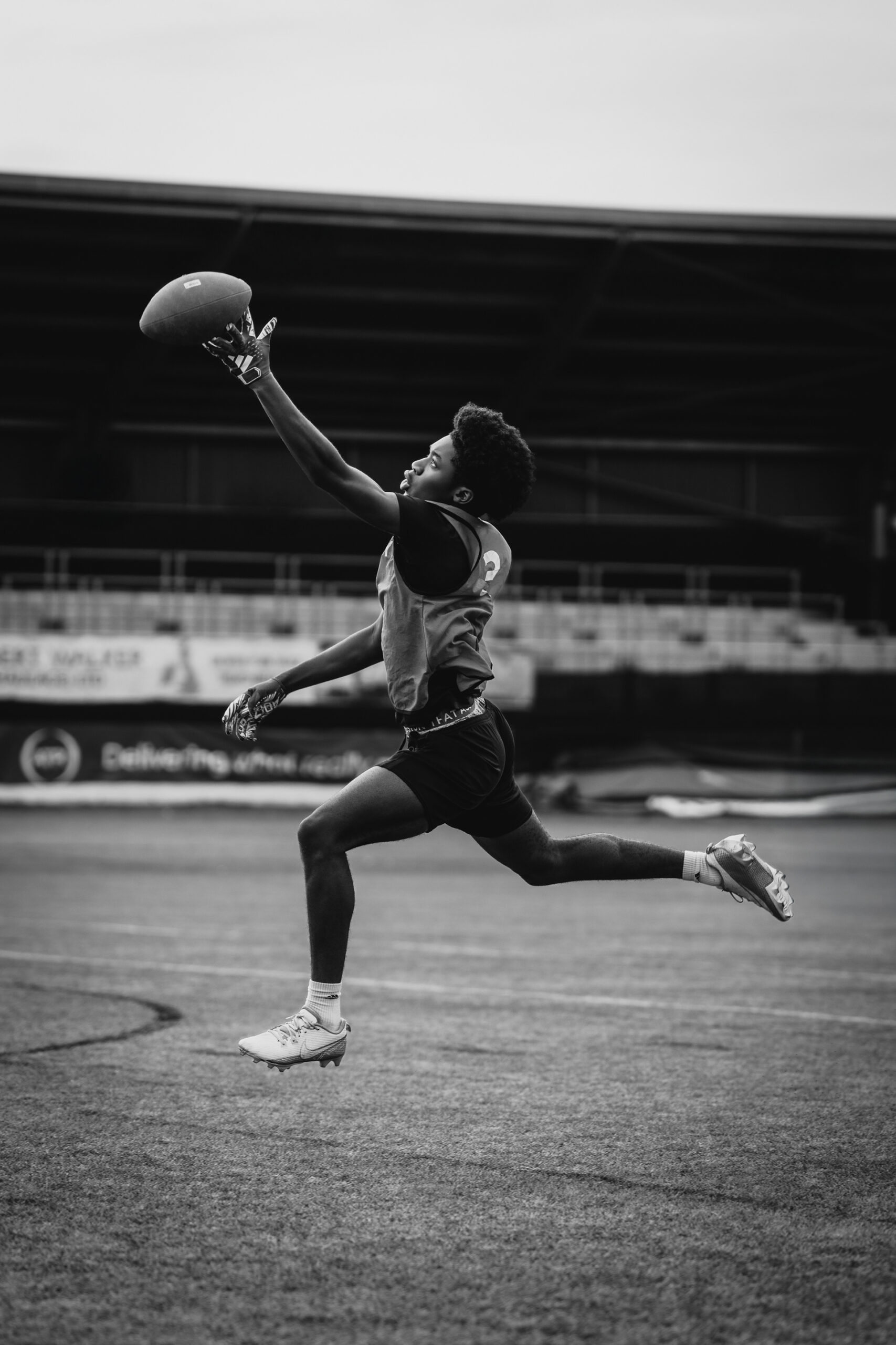 Sports & Fitness Photography | A young athlete in a sports vest leaps mid-air, fully outstretched with one arm reaching for a football. The background shows empty stadium seats and a covered structure. The image is black and white, emphasising motion and effort. Nathan Mountain | Hampshire & London Photography | Hampshire & London Camera Man