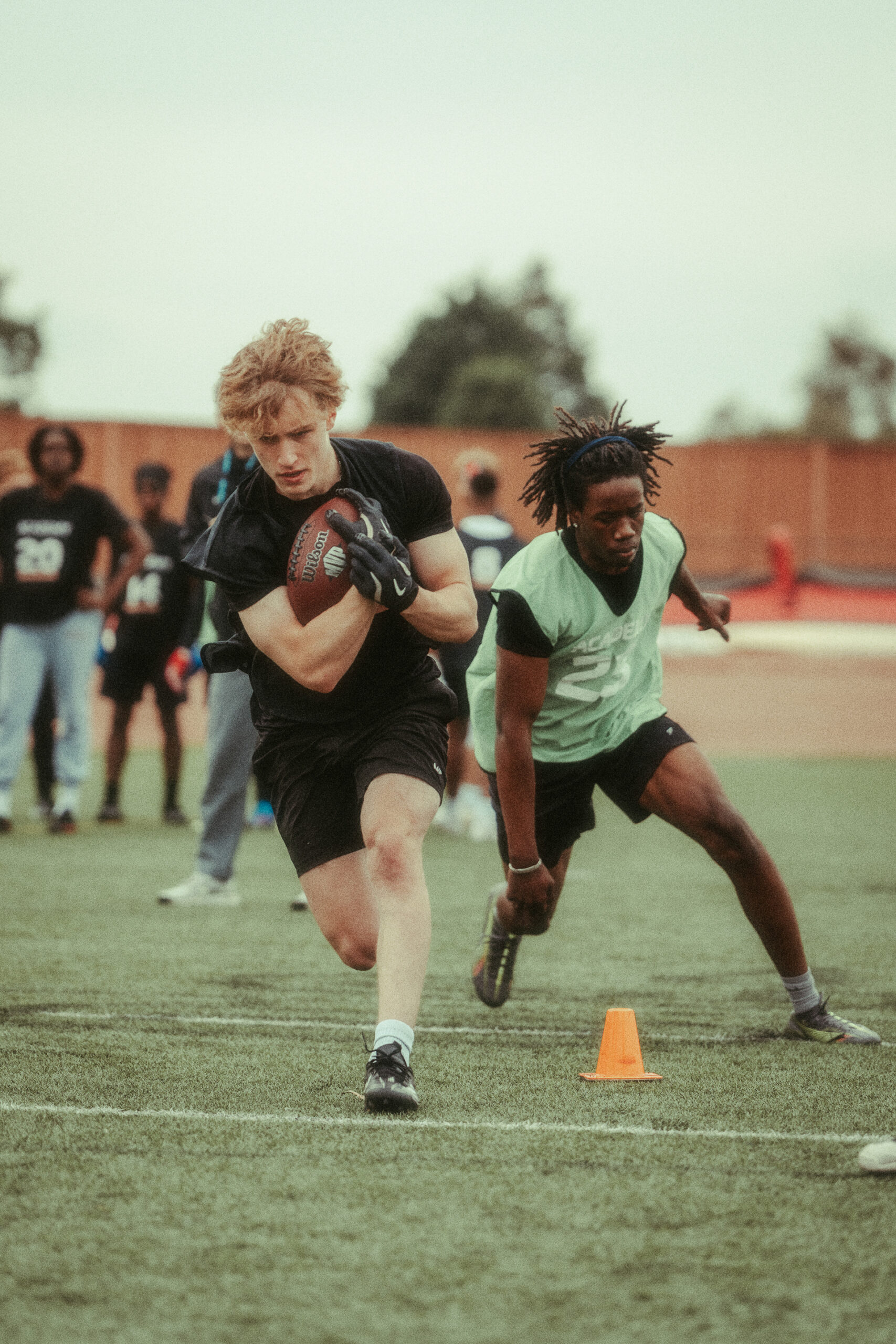 Sports & Fitness Photography | A young man with light skin and red hair runs with a football, dodging an orange cone, whilst another player in a green vest chases him on a grassy pitch. Several people and a fence are visible in the blurred background. Nathan Mountain | Hampshire & London Freelance Photographer | Hampshire & London Camera Man