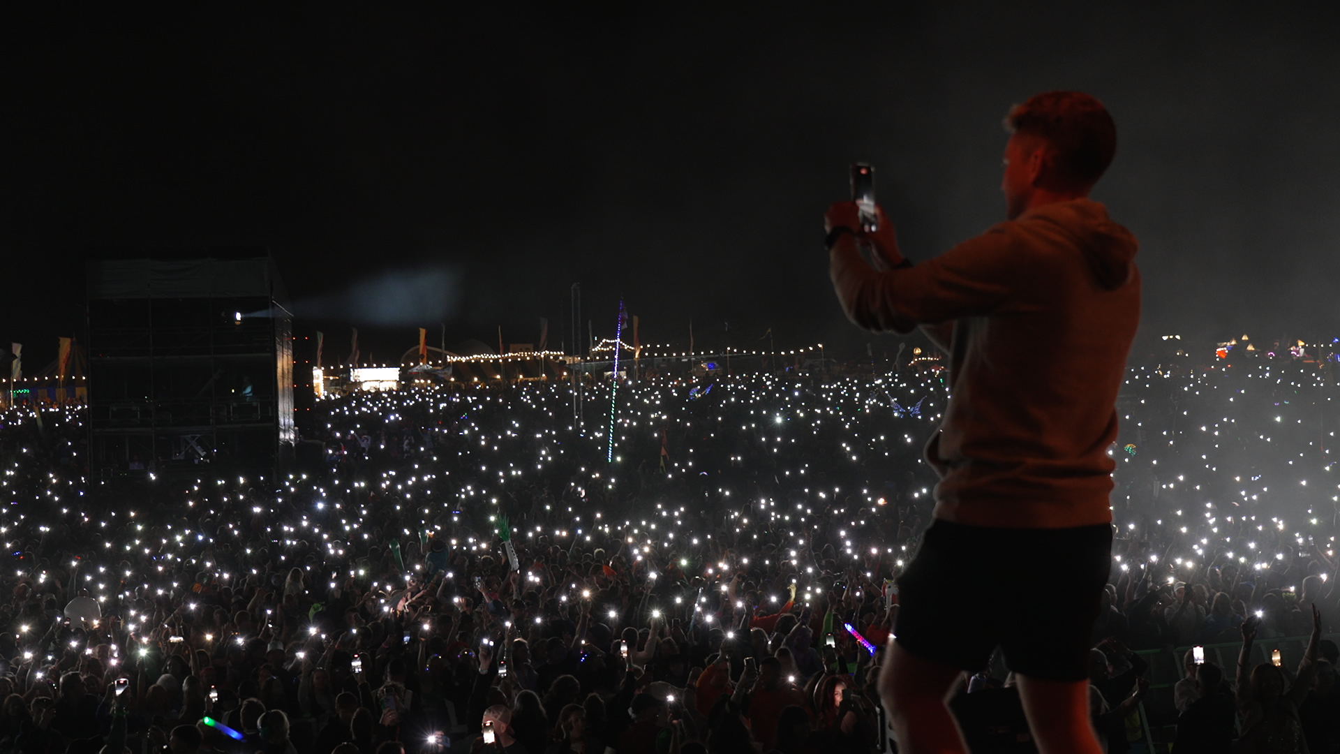 Event Photography | A person stands on stage at night, facing a large outdoor crowd. The audience hold up phones, creating a sea of small white lights. Colourful festival lights and tents are visible in the background, adding to the lively atmosphere. Nathan Mountain | Hampshire & London Freelance Photographer | Hampshire & London Camera Man
