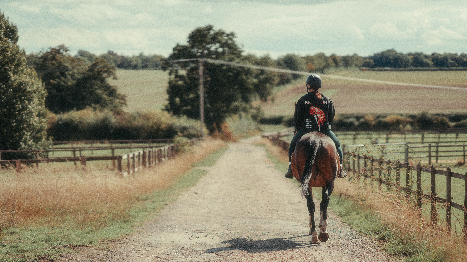 Pet Photography | A person wearing a black helmet and dark jacket rides a brown horse down a rural dirt road, flanked by wooden fences and grassy fields, with trees and cloudy sky in the background. The rider and horse are seen from behind. Nathan Mountain | Hampshire & London Freelance Photographer | Hampshire & London Camera Man